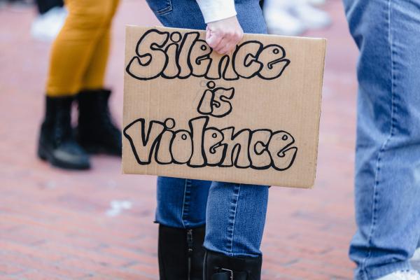 woman holds a cardboard sign which has the words 'silence is violence'